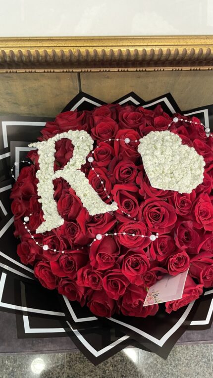 Red roses with a white floral initial and heart, decorative bead accents, black wrapping and a visible banknote.