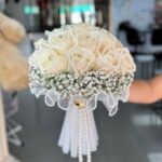Ivory roses above baby’s breath with a scalloped bead-style collar and hanging decorative strands, shown in a supplied reference photograph.