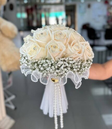 Ivory roses above baby’s breath with a scalloped bead-style collar and hanging decorative strands, shown in a supplied reference photograph.