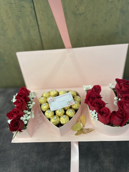 Red roses, wrapped chocolates and a banknote presentation arranged inside a pale pink letter-style gift box.