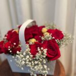 Red roses, white baby’s breath and gold-wrapped chocolates in a handled pale gift basket.
