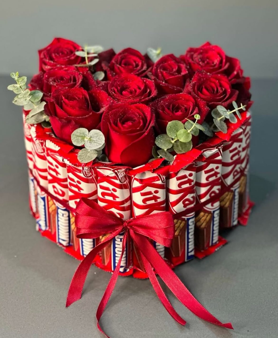 Red roses and greenery in a round presentation surrounded by wrapped chocolate bars and red ribbon, shown in a supplied reference image. Red roses and greenery in a round presentation surrounded by wrapped chocolate bars and red ribbon, shown in a supplied reference image.