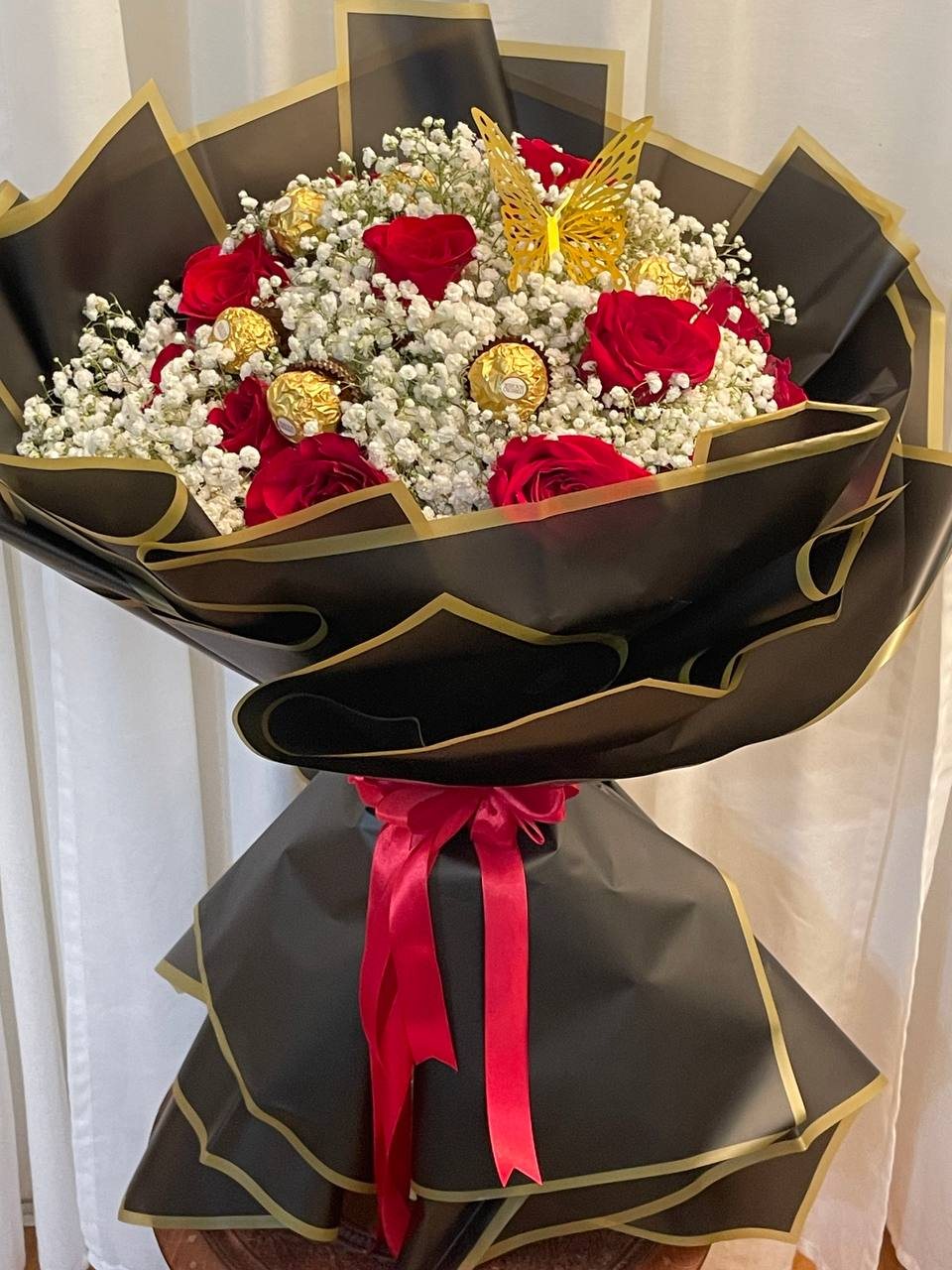 Red roses and gold-wrapped chocolates set among abundant white baby’s breath, with black-and-gold wrapping and butterfly decoration. Red roses and gold-wrapped chocolates set among abundant white baby’s breath, with black-and-gold wrapping and butterfly decoration.