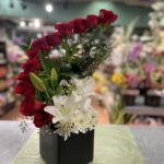 An asymmetric arc of red roses above white lilies and foliage in a black box.