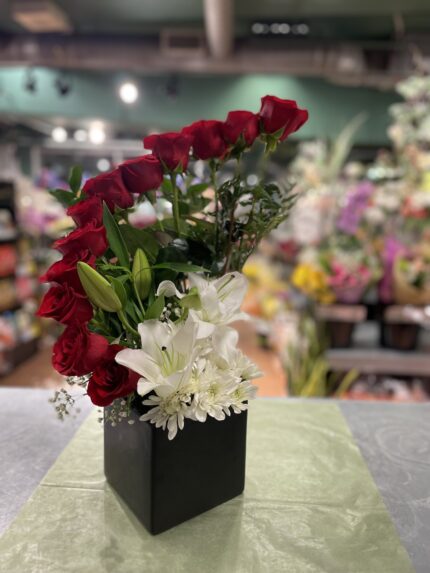 An asymmetric arc of red roses above white lilies and foliage in a black box.