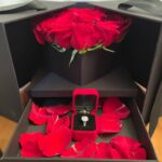 Red roses in a black presentation box with a petal-lined drawer displaying a jewellery case and jewellery.