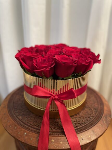 A rounded red rose arrangement in a ribbed gold-tone hatbox, finished with red ribbon.