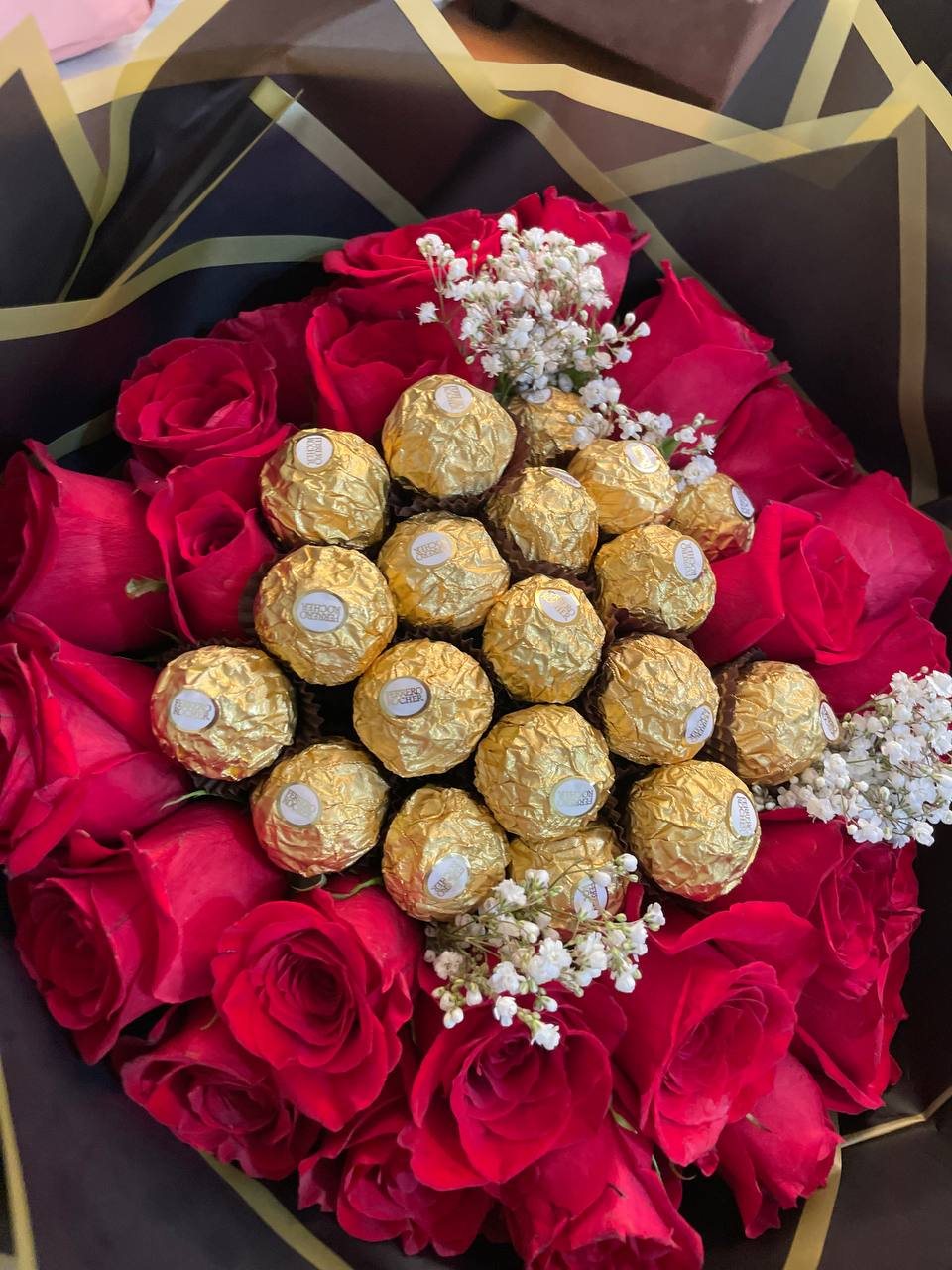 A red rose bouquet surrounding a centre of gold-wrapped chocolates with white baby’s breath and black-and-gold wrapping. A red rose bouquet surrounding a centre of gold-wrapped chocolates with white baby’s breath and black-and-gold wrapping.