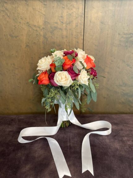 Orange, red and ivory roses with flowing green foliage and a trailing white ribbon.