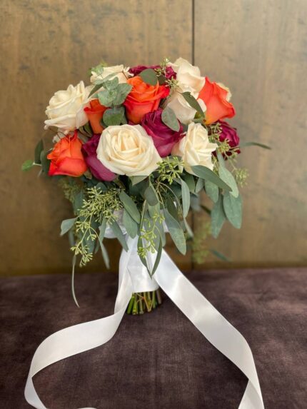 Orange, red and ivory roses with flowing green foliage and a trailing white ribbon.
