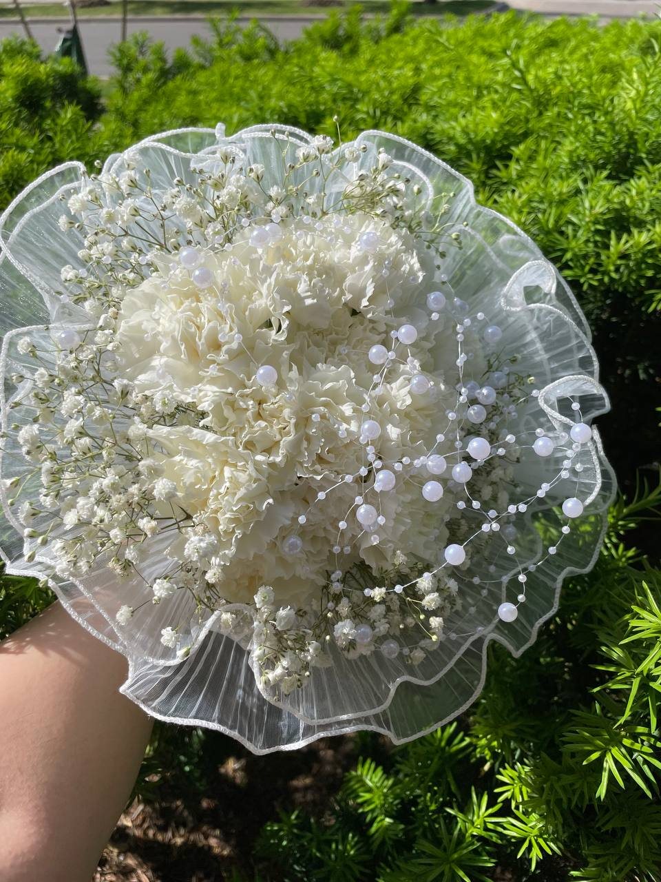 White carnations and baby’s breath inside a ruffled translucent collar with decorative white accents. White carnations and baby’s breath inside a ruffled translucent collar with decorative white accents.