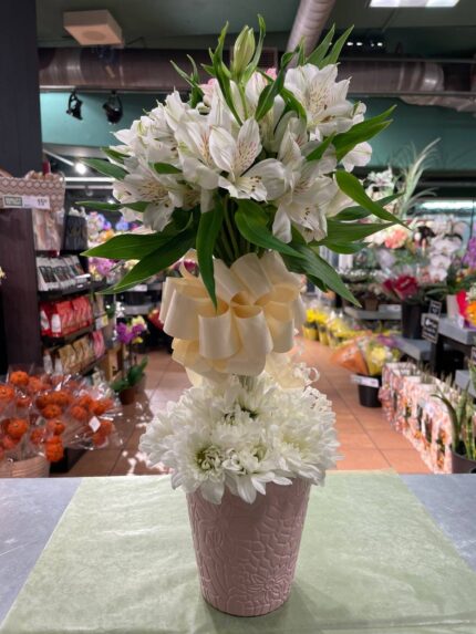 A tall white floral arrangement with lilies, airy white blooms and a ribboned vertical design in a textured container.