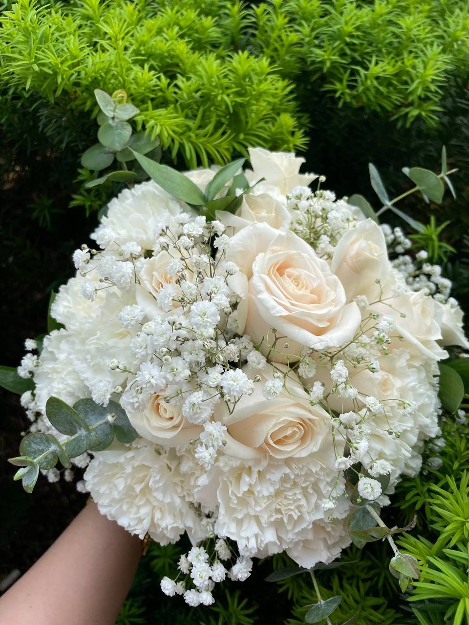 White roses and ruffled white flowers mixed with baby’s breath and eucalyptus-style greenery in a rounded bridal bouquet.