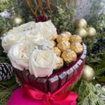 White roses and gold-wrapped chocolates in a round box surrounded by wrapped chocolate bars and a red ribbon.